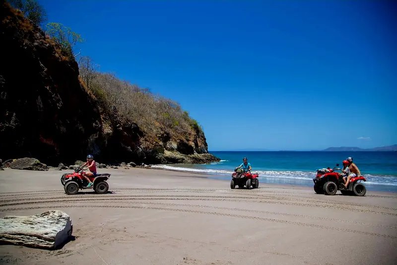 Three guests riding red ATVs on a quiet beach beside a rocky headland in Guanacaste, Costa Rica